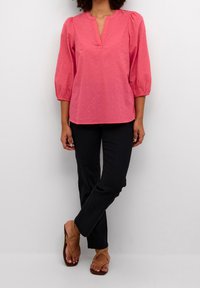 Donna che indossa una blusa corallo rosa con maniche a tre quarti, pantaloni neri e sandali con cinturini incrociati color sabbia, in piedi su uno sfondo bianco.