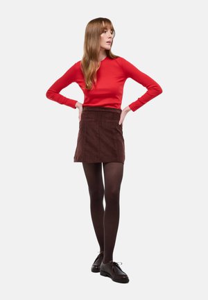 Haut rouge à manches longues associé à une mini-jupe en velours côtelé marron. Le mannequin porte des collants foncés et des chaussures noires à lacets, les bras sur les hanches.