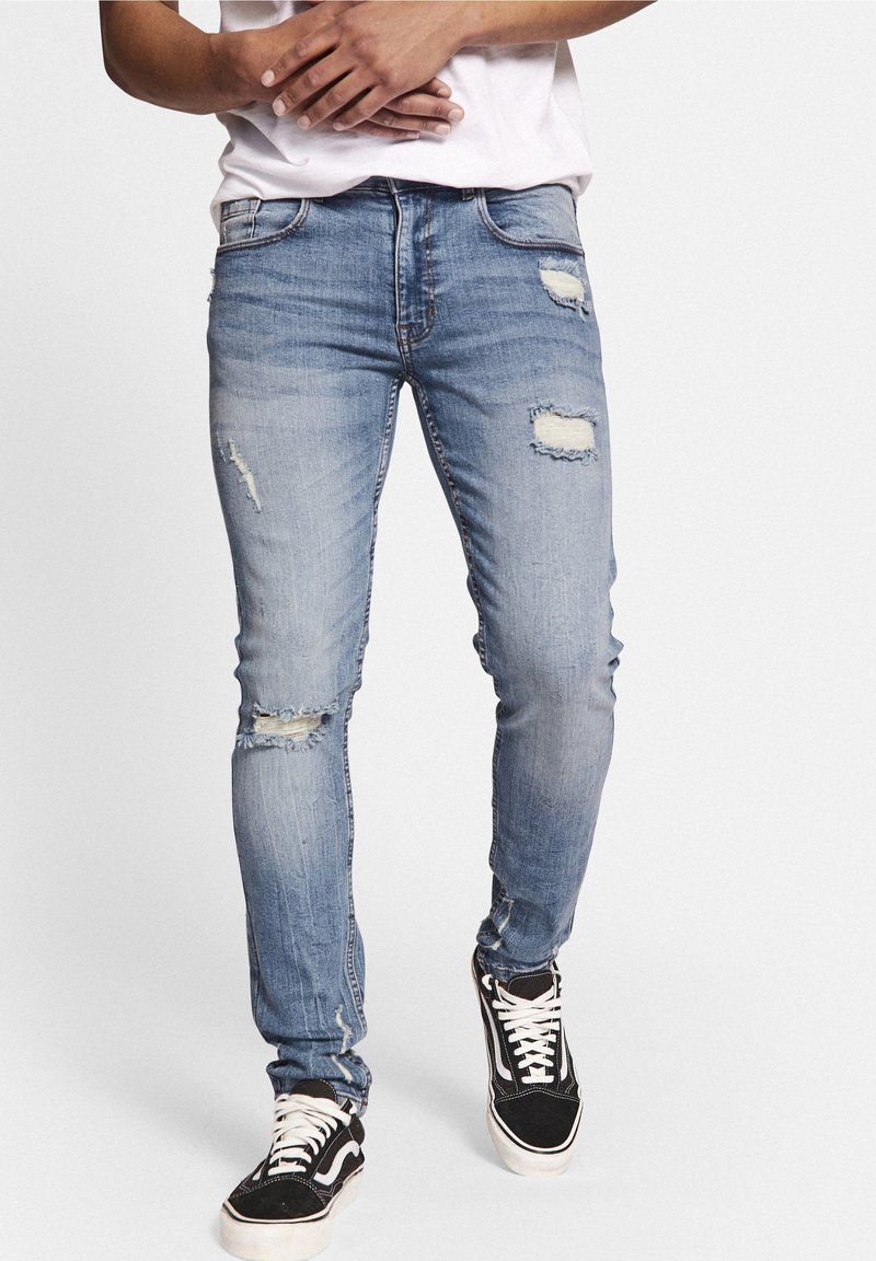 Jeans de mezclilla azul claro con un ajuste slim, áreas desgastadas y múltiples bolsillos. Combinados con zapatillas negras y blancas.