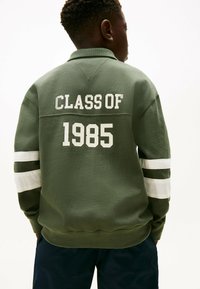 Groene sweatshirt met witte tekst "CLASS OF 1985" op de achterkant, met witte en groene strepen op de mouwen en een kraag.