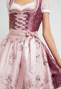 Robe dirndl avec un corsage rose foncé à motifs et des finitions en dentelle, un détail de corset en ruban satiné, et une jupe rose clair ornée de broderies florales.