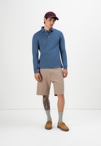 Mann in blauem Langarm-Polo, beigen Shorts, grauen Socken, braunen Schuhen und burgunderfarbener Kappe, stehend mit sichtbarem Tattoo am linken Bein vor weißem Hintergrund.