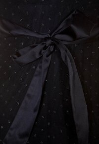 Tissu noir à motif à pois, agrémenté d'un large nœud en satin noué à la taille. Le matériau présente un brillant subtil et une texture lisse.