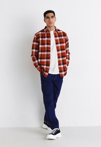 Jeune homme debout contre un mur blanc portant une chemise à carreaux rouges, un t-shirt blanc, un pantalon bleu foncé et des baskets noir et blanc.