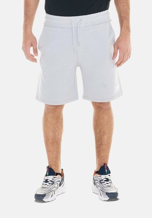 Uomo che indossa pantaloncini grigio chiaro con coulisse, maglietta nera e scarpe sportive bianche con dettagli neri e arancioni, in piedi su uno sfondo bianco.