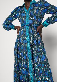 Abito lungo a fantasia floreale con base nera e fiori blu e bianchi, colletto teal e cintura coordinata, realizzato in un tessuto morbido.