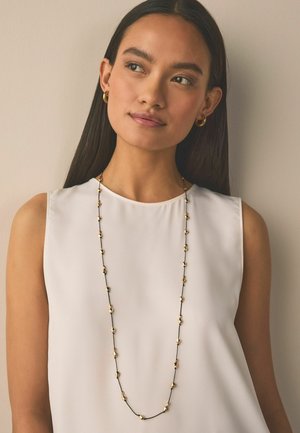 Goldener Anhänger an einer schwarzen Schnur mit abwechselnden goldenen Formen. Das Modell trägt ein ärmelloses weißes Top vor neutralem Hintergrund.