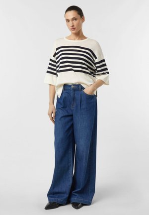 Donna in piedi che indossa un maglione ampio a righe bianche e nere, jeans blu a gamba larga e scarpe nere con punta, su uno sfondo neutro.