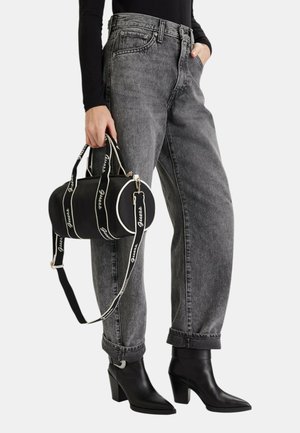 Persona che indossa jeans grigi a vita alta e stivaletti neri, che tiene in una mano una borsa cilindrica nera con cinturini brandizzati.