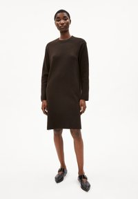 Braun gestricktes Kleid mit langen Ärmeln, Rundhalsausschnitt und seitlichen Schlitzen am Saum. Kombiniert mit schwarzen flachen Schuhen mit Schnallelementen.