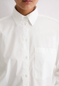 Eine Person trägt ein frisches weißes Hemd mit Knöpfen, spitzem Kragen und einer Brusttasche, und steht vor einem hellen Hintergrund.