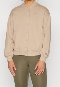 Giacca felpata beige con chiusura a bottoni, polsini e orlo a coste. Realizzata in tessuto morbido, presenta un piccolo logo sulla manica sinistra.
