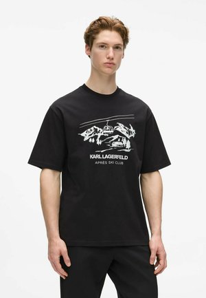 Junger Mann mit kurzen hellbraunen Haaren, der ein schwarzes Karl Lagerfeld T-Shirt mit weißem Ski-Berg- und Seilbahndesign trägt.