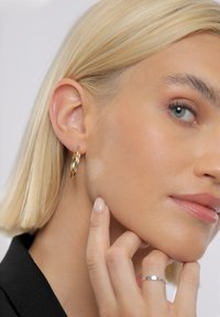 Auksinis lankelio formos auskaras su poliruotu paviršiumi, dėvimas trumpais blondinų plaukais. Matoma lygi odos tekstūra ir neutralių atspalvių nagų lakas.