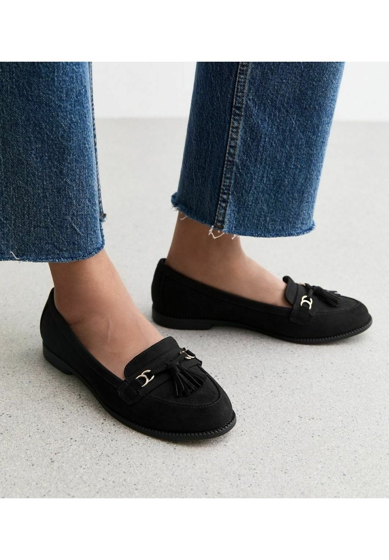 Mocassins en daim noirs avec un détail à pampille et des accents en or ; petit talon ; associés à un pantalon en denim bleu taille courte.