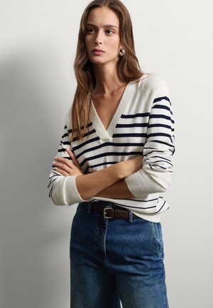 Junge Frau mit langen braunen Haaren, die einen weißen und dunkelblau gestreiften Pullover und blaue Jeans trägt, steht mit verschränkten Armen vor einer schlichten Wand.