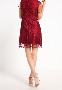 Rotes Spitzenkleid mit durchsichtigen Ärmeln, knielang und mit floralen Mustern. Getragen mit hautfarbenen High Heels, die über Knöchelriemen verfügen.