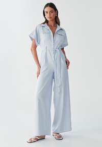 Mono azul claro de manga corta con cuello camisero, cierre frontal con botones, dos bolsillos en el pecho y cintura anudada. Corte de pierna ancha y textura suave.