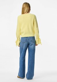 Blond kvinna stående vänd bort, klädd i en gul tröja med knytna ärmar och vida blå jeans mot en enfärgad vit bakgrund.