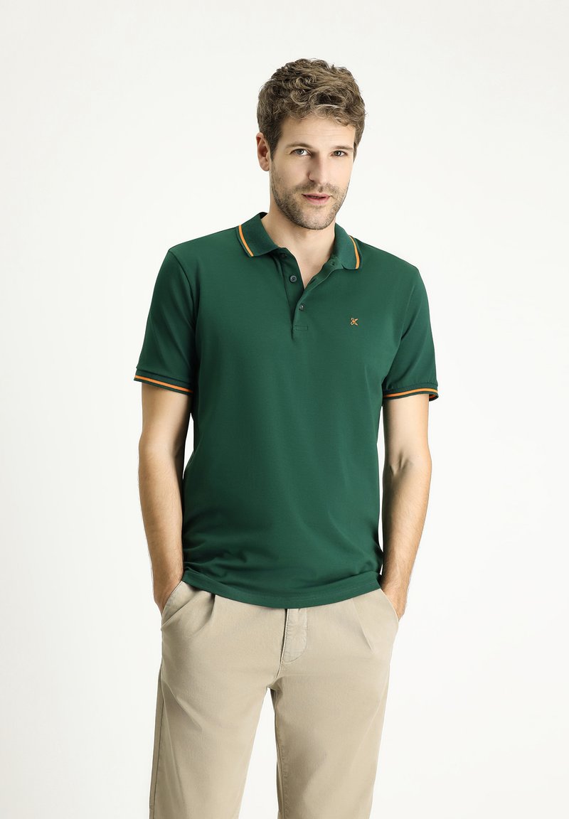 Polo shirt verde scuro con dettagli arancioni sul colletto e sulle maniche, realizzato in un tessuto liscio. Indossato con pantaloni beige chiaro.