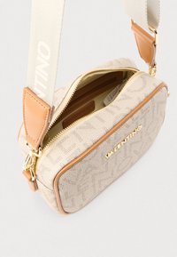 Beżowa torba o fakturze, z brązowymi skórzanymi akcentami i złotymi elementami metalowymi. Posiada górny suwak i szeroki, regulowany pasek z napisami.