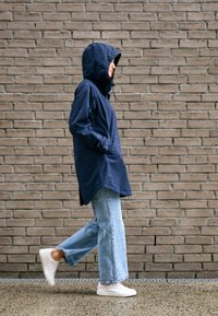 Osoba kráčajúca popri tehlovej stene v tmavomodrom nepremokavom kabáte s kapucňou, svetlomodrých džínsoch a bielych teniskách, ruky v kapsách.