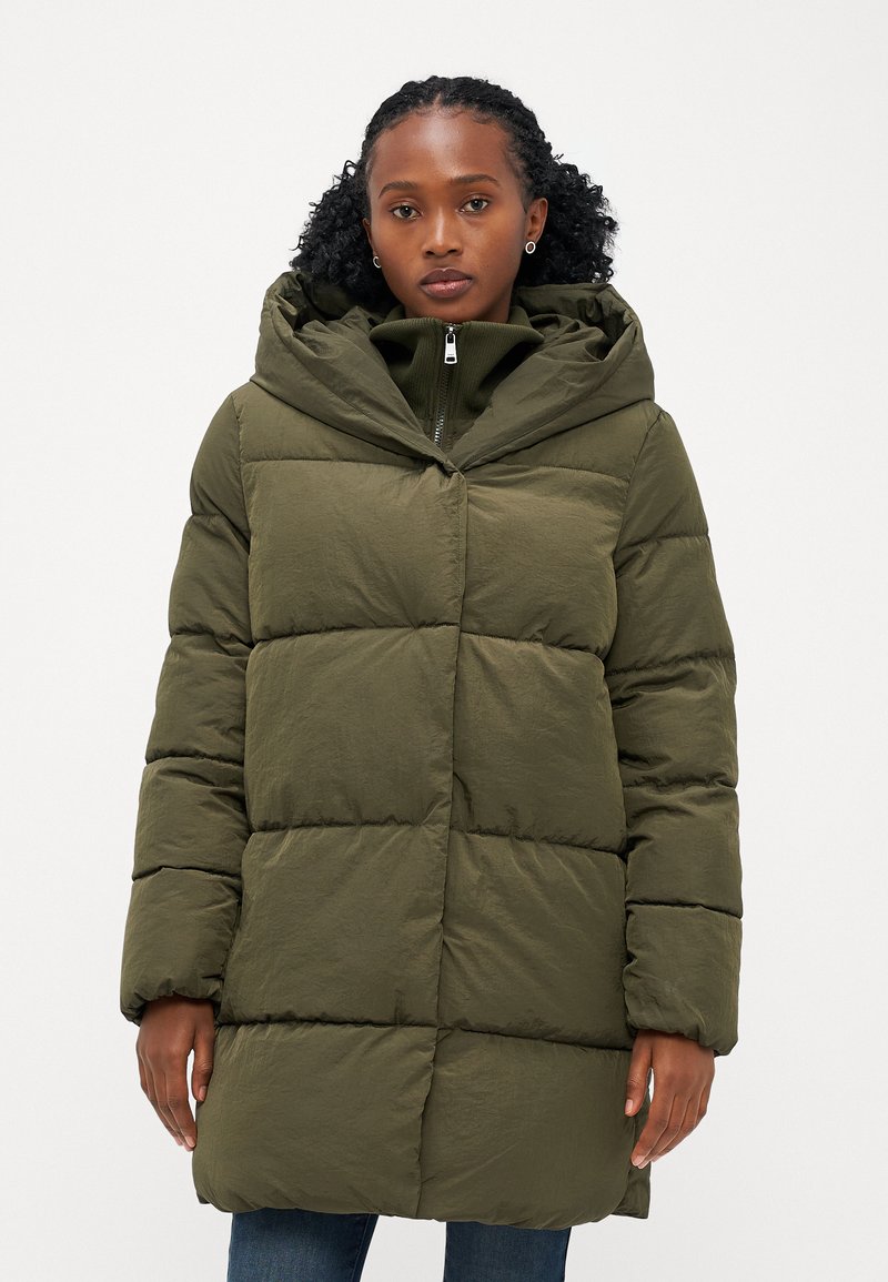 Doudoune vert olive avec une grande capuche, fermeture éclair et forme carrée. Présente un design matelassé avec des coutures visibles et un tissu texturé.