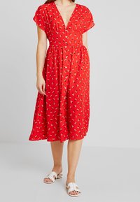 Vestido rojo con flores, con un escote en V profundo, mangas cortas y un corte relajado, que presenta pequeños patrones de flores blancas y negras y una falda de longitud media.