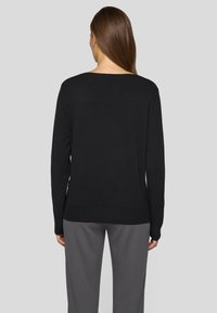 Schwarzer Pullover mit Rundhalsausschnitt und geripptem Saum, ausgestattet mit langen Ärmeln. Über grauen Hosen getragen, zeigt er eine glatte Textur und ein schlichtes Design.