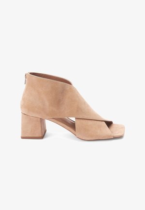 Bottines à talon bloc en daim beige, ouvertes au niveau des orteils, avec un design asymétrique et une fermeture éclair à l'arrière.