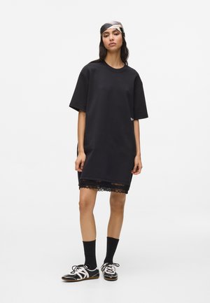 Frau trägt ein schwarzes, übergroßes T-Shirt-Kleid mit Spitzenbesatz, schwarze Socken, schwarz-weiße Turnschuhe und ein weiß-schwarzes, gemustertes Kopftuch.