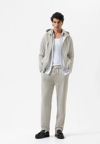 Man in een lichtgrijze hoodie, gestreepte overhemden, witte tanktop, bijpassende sweatpants en zwarte loafers, staande tegen een witte achtergrond.