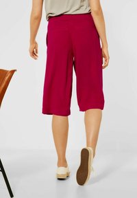 Magenta culottes dengan potongan longgar, pinggiran datar, saku samping, dan saku belakang, dipadukan dengan sepatu wedge berwarna terang.