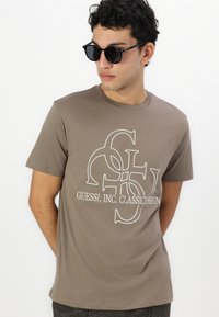 Brown cotton t-shirt featuring white logo print "GUESS?, INC. CLASSIC DESIGN." Crew neck and short sleeves. Model wearing sunglasses.