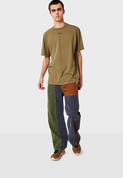Ung man som bär en olivgrön oversized T-shirt och färgblockerade lapptäcktsjeans, går framåt framför en enfärgad bakgrund.