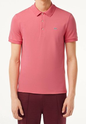Różowa koszulka polo wykonana z miękkiego materiału, z kołnierzykiem, trzema guzikami i małym haftowanym logo na lewej piersi.