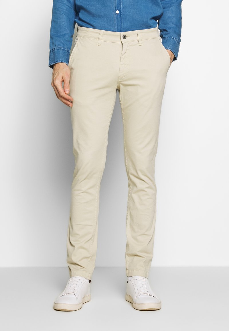 Beige Baumwoll-Chinos im Slim-Fit-Stil, mit Fronttaschen und Knopfverschluss, kombiniert mit weißen Sneakers.