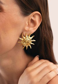 Orecchini d'oro a forma di sole con superfici testurizzate e spuntoni allungati, indossati all'orecchio. Piccole protuberanze aggiungono dettagli al pezzo centrale.