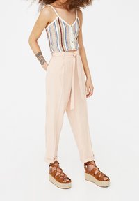 Lichtroze hoge taille broeken met een knooptaille en plooien. Gecombineerd met een kleurrijke gestreepte tanktop en bruine sandaaltjes met plateauzolen.