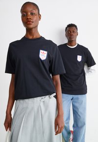 Melnas īsām piedurknēm t-krekli ar sarkano un zilo Adidas logo uzšišanu, ar klasisku piegriezumu. Viens modelis valkā pelēku svārku ar krokām.