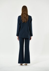 Completo su misura blu navy con giacca a un petto e pantaloni svasati, caratterizzato da un tessuto liscio e dettagli con bottoni sulle maniche.