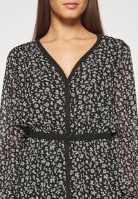 Femme portant une robe noire boutonnée avec un petit motif floral blanc, décolleté en V, manches longues transparentes et taille cintrée.