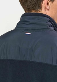 Homme portant une veste bleu foncé avec un petit emblème de drapeau à rayures horizontales rouges, blanches et bleues sur le haut du dos près du col.