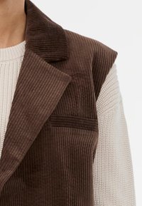 Brązowa kamizelka z korduroju z szerokim kołnierzem, z pionowymi paskami i kieszenią naszywaną, nałożona na strukturalny beżowy sweter.