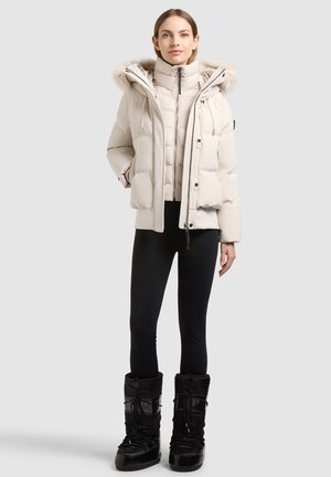 Femme portant une doudoune beige avec capuche en fourrure, des leggings noirs et des bottes d’hiver noires, debout devant un fond clair uni.