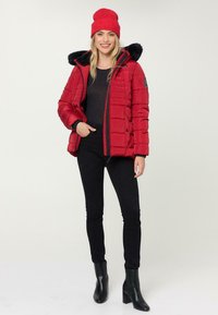Rote Daunenjacke mit schwarzem Kunstfellfutter in der Kapuze, Reißverschluss und horizontalem Steppmuster. Schwarze enganliegende Hose und schwarze Stiefeletten.