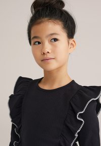 Top nero a coste con maniche arricciate bordate di bianco, scollatura rotonda e texture morbida, che mostra un primo piano dei dettagli del design.