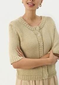 Cardigan à manches courtes tricoté en beige clair avec un col et un ourlet côtelés, comportant quatre grands boutons à l'avant. Tissu texturé.
