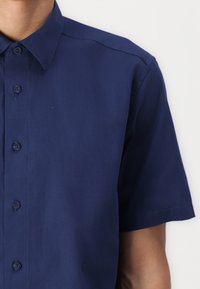 Camisa de manga corta en color navy, hecha de algodón. Presenta un cierre de botones en la parte delantera, cuello puntiagudo y una textura suave y uniforme con detalles de costura sutiles.