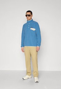 Pulôver azul de lã com bolso frontal bege e gola com botões, combinado com calças bege e ténis brancos, complementado por grandes óculos de sol.